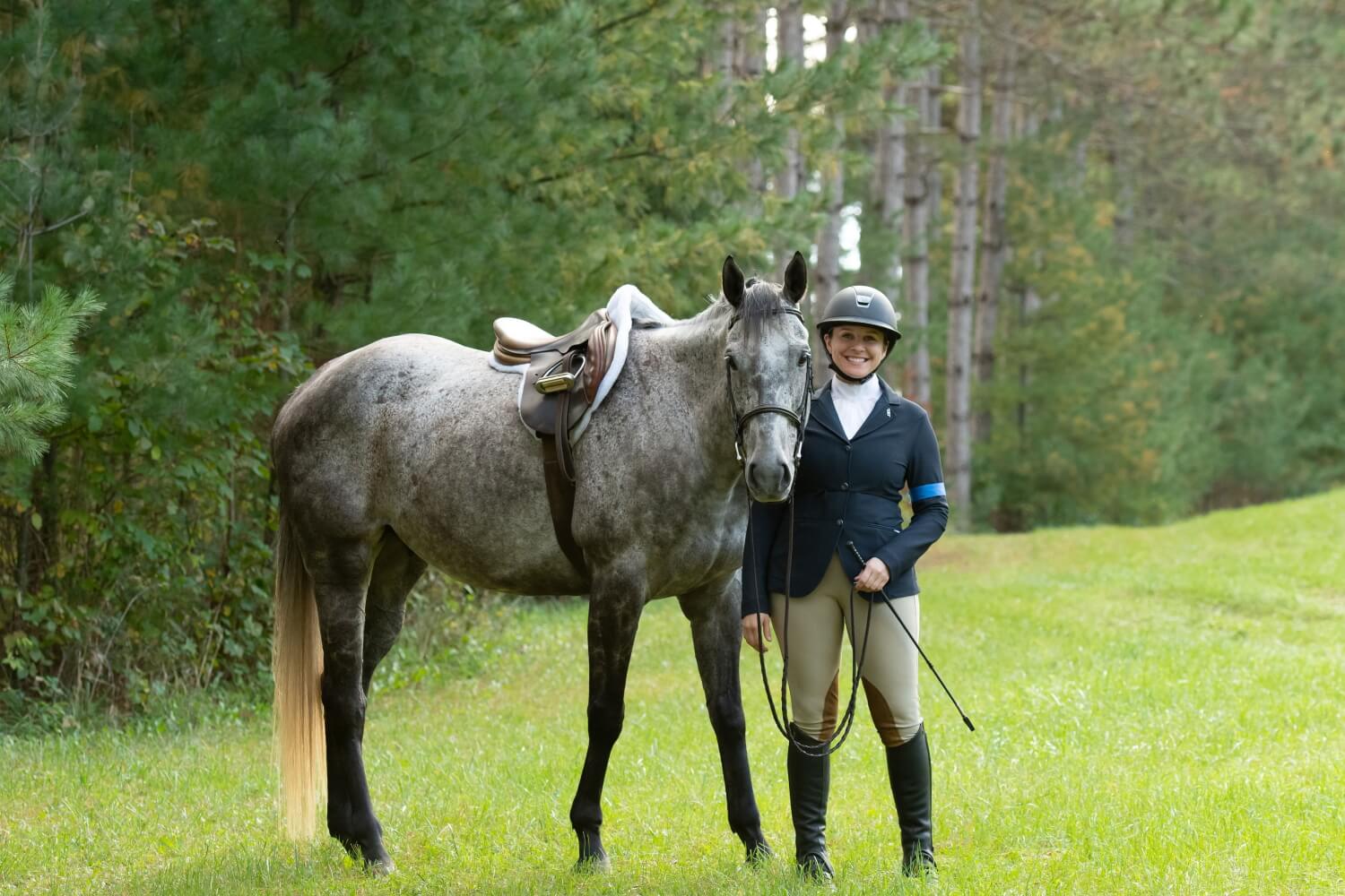 Rider in traditional English uniform posing by a Thoroughbred horse in a forest clearing